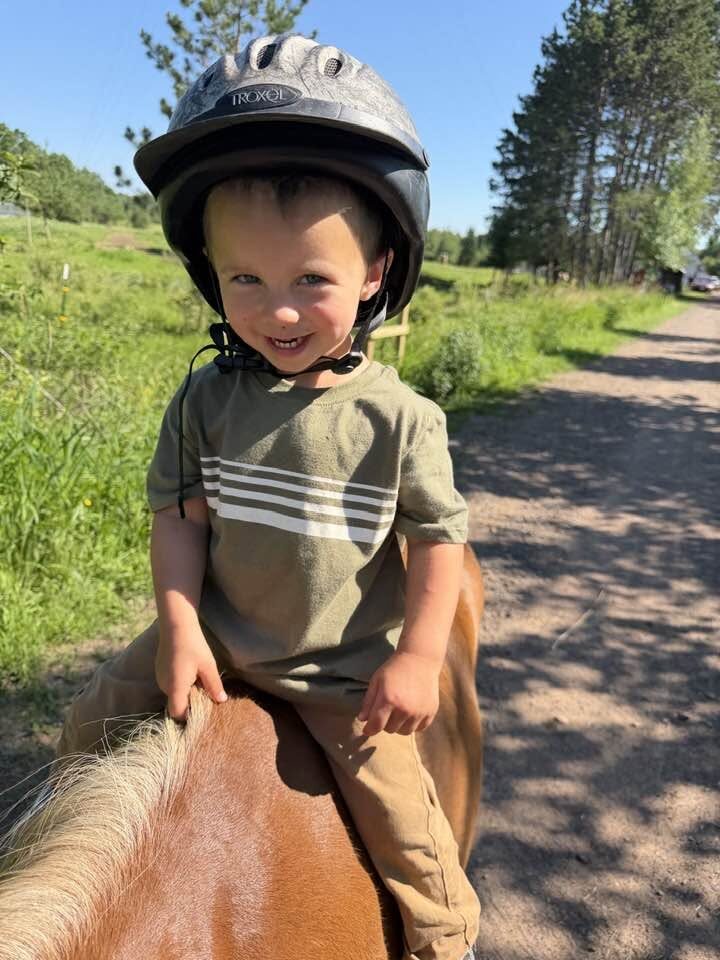 A laughing child in a riding helmet sits on a pony under a blue sky at Full Circle Vaulting