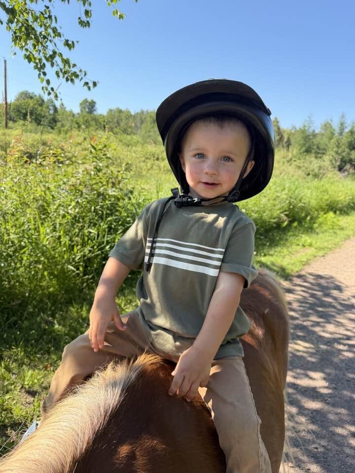 A child in a riding helmet rides a pony along a lush green trail at Full Circle Vaulting