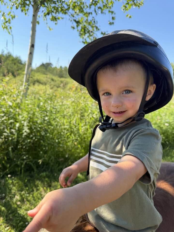 A young child in a helmet rides a pony past birch trees and tall grass